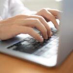 ノートパソコンを操作している人。明るいナチュラルな木製のデスクで白いシャツを着た人がPCのキーボードをタイピングしている。